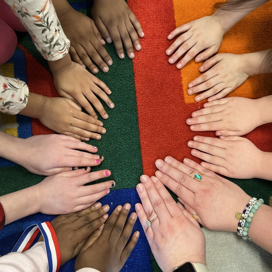 Un grupo de manos de todo tipo, dispuestas en círculo sobre una alfombra de colores, en las que se aprecian diferentes tonos de piel, anillos y pulseras.